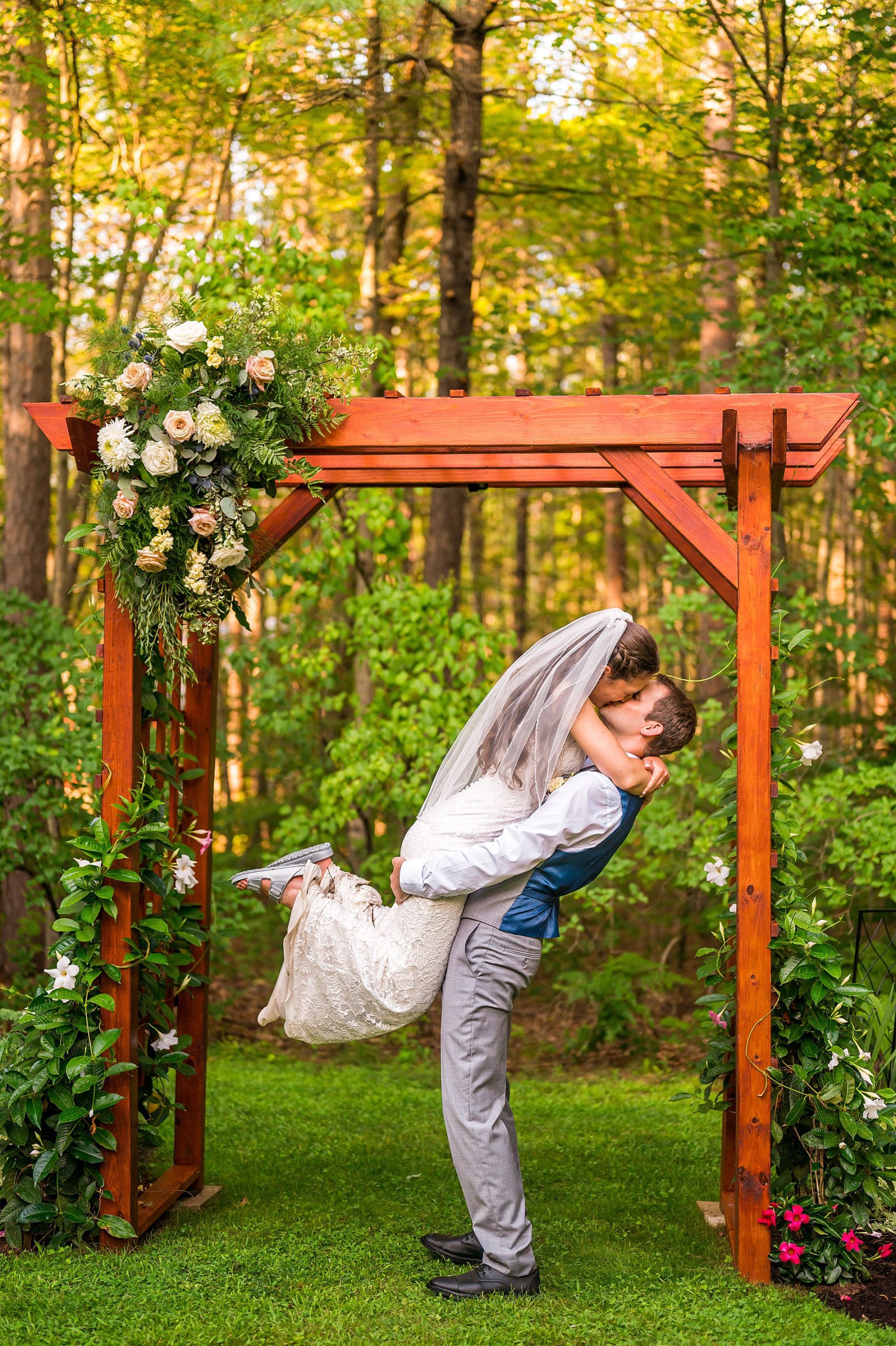groom lifts bride up under wooden arbor at intimate summer wedding 