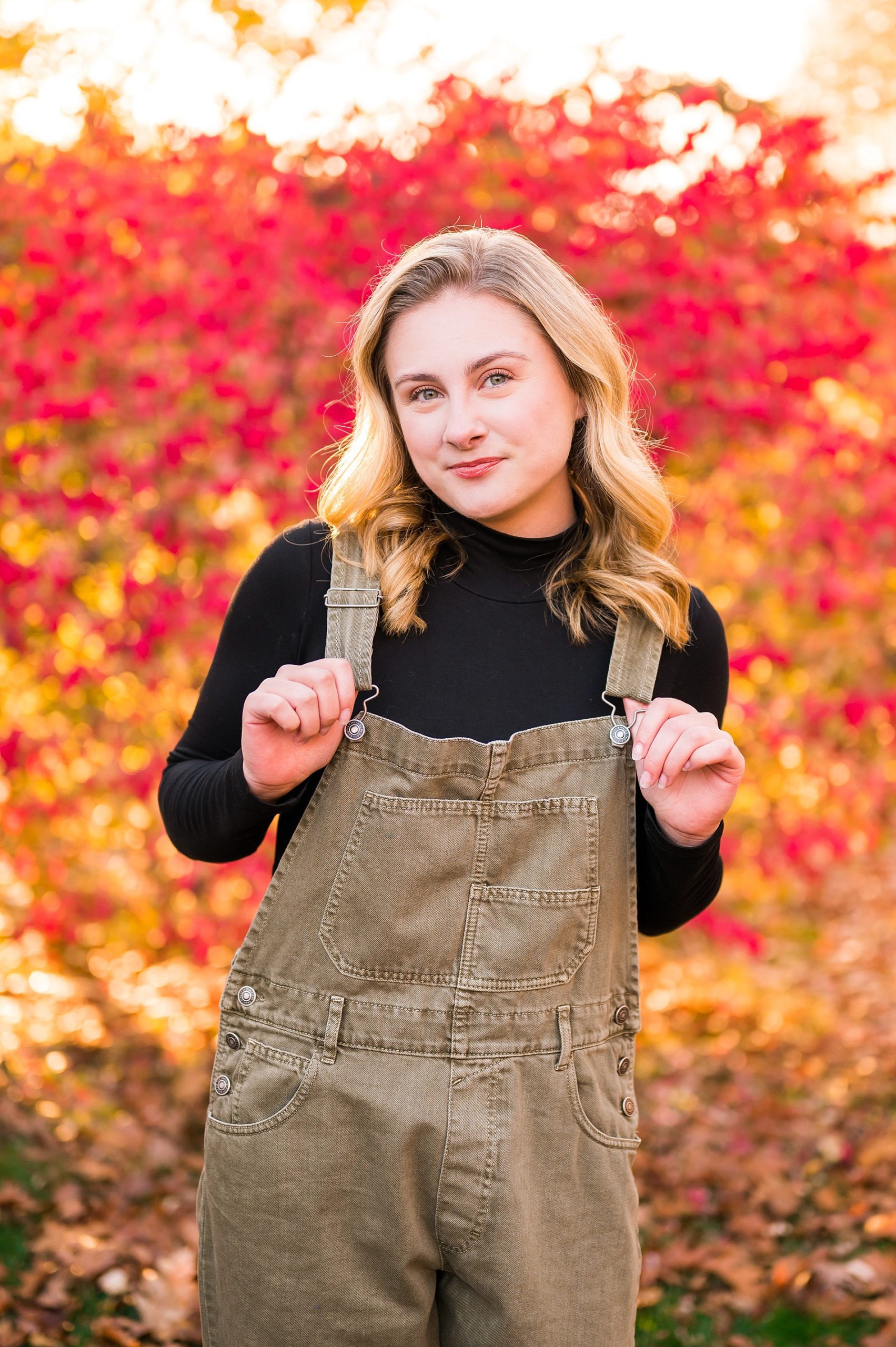 senior girl wearing tan overalls with red fall foliage in the background 