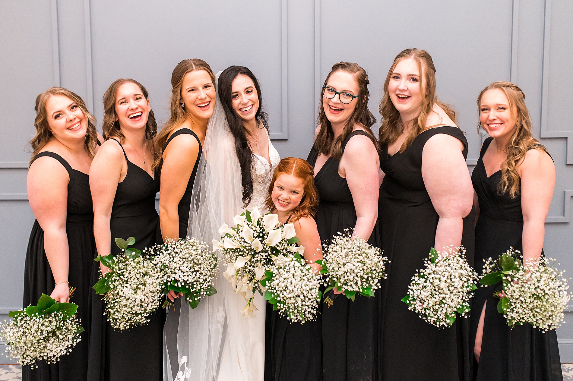 bride and bridesmaids in black dresses hold white wedding bouquets of babies breath